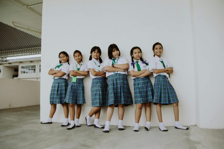 A group of six smiling schoolgirls in uniform standing indoors with arms crossed, displaying friendship.