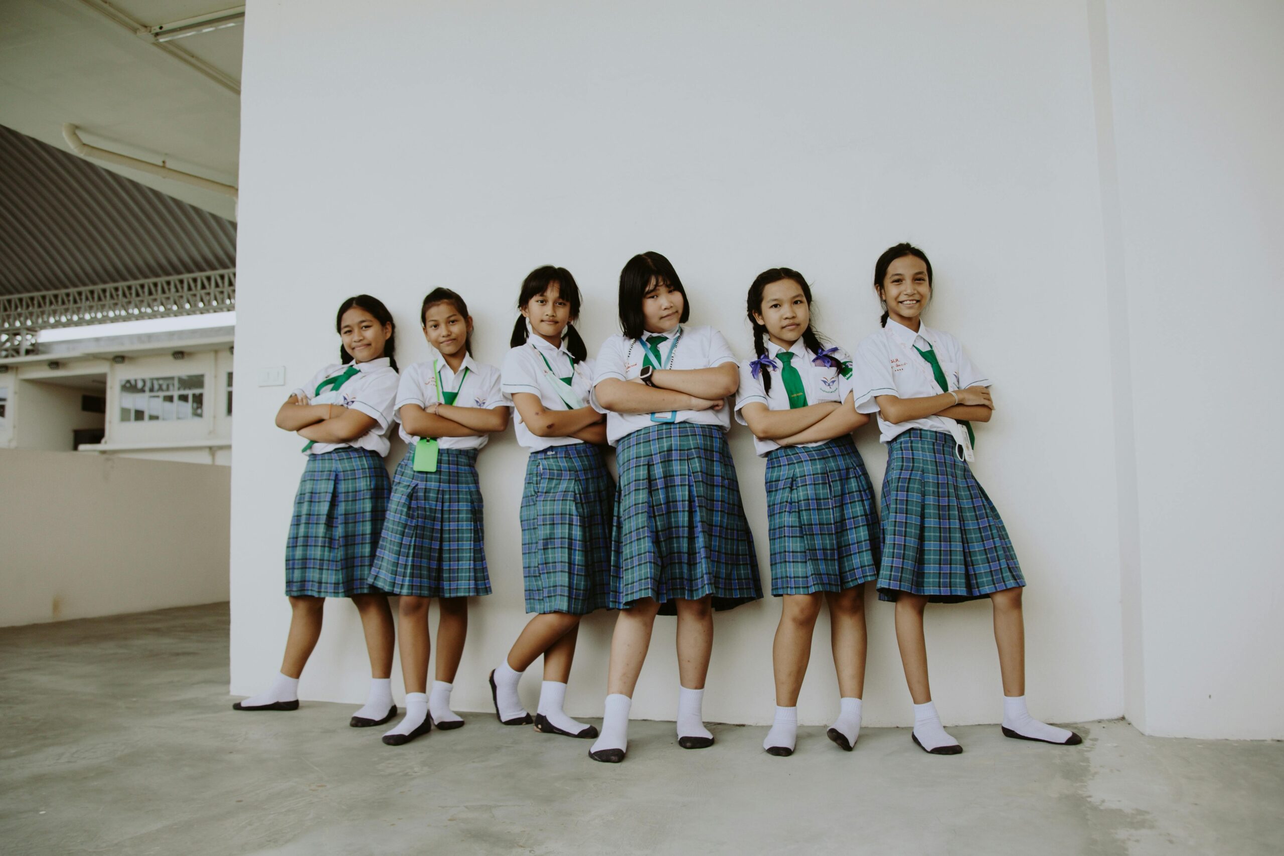 A group of six smiling schoolgirls in uniform standing indoors with arms crossed, displaying friendship.